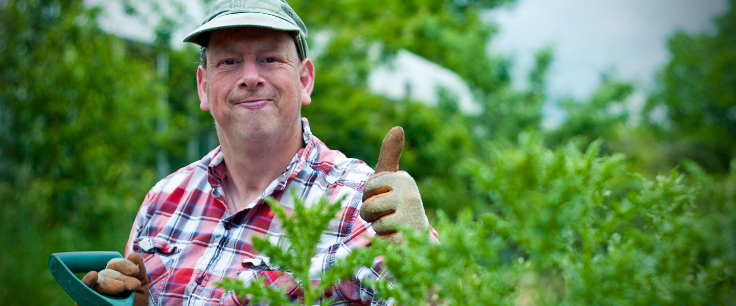 An Earthworker standing amongst bushes with his thumbs up, wearing gardening gloves, holding the handle to a shovel