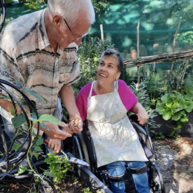 An Earthworker in a wheelchair holding a hand shovel over a raised planting bed, smiling at a volunteer