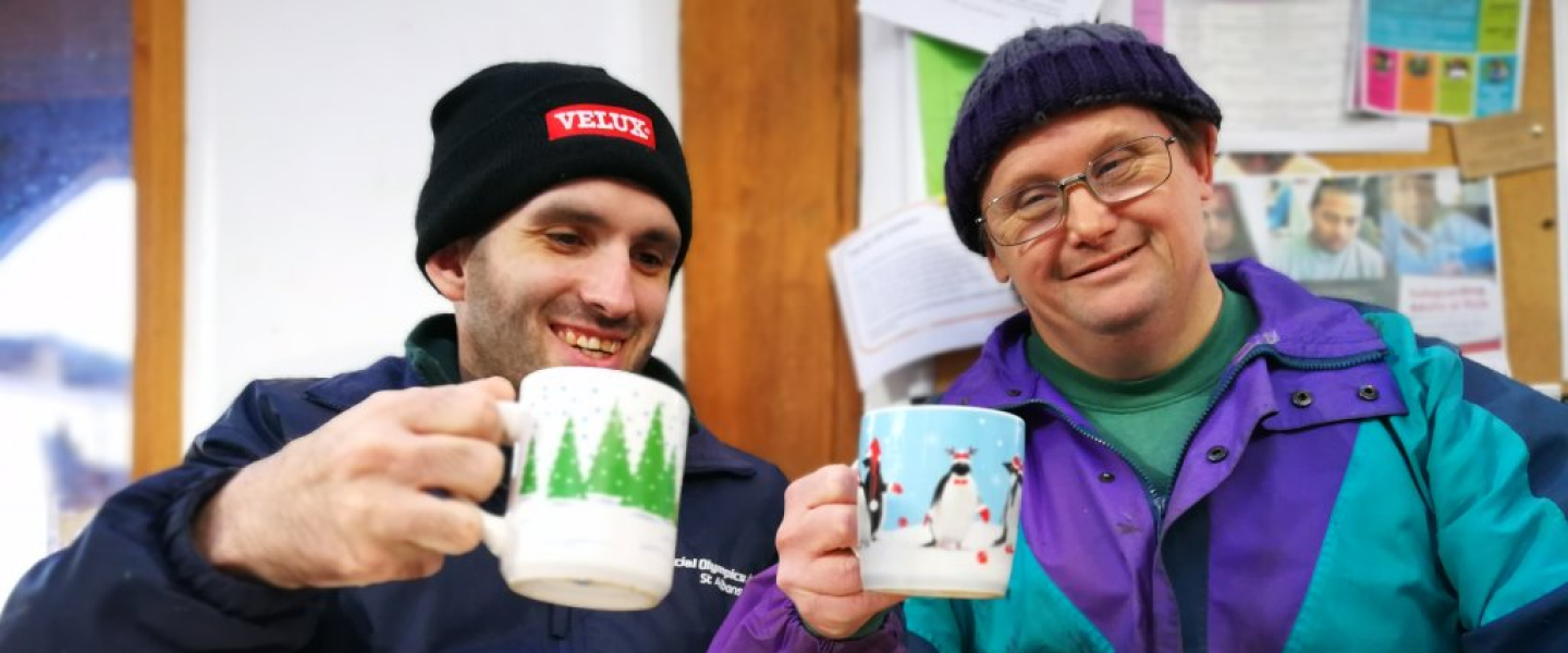 Two Earthworkers sat together holding Christmas-themed mugs, wearing beanie hats and coats