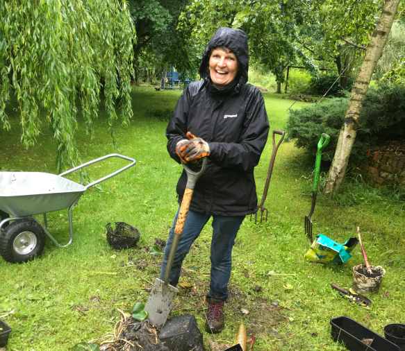 A volunteer wearing a raincoat and gardening gloves in the gardens, holding a shovel and surrounded by gardening tools
