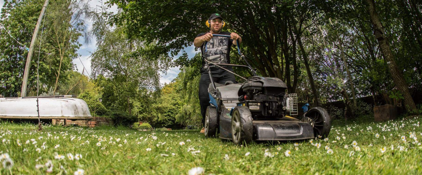 A volunteer cutting the grass with a lawn mower in the gardens, wearing ear defenders