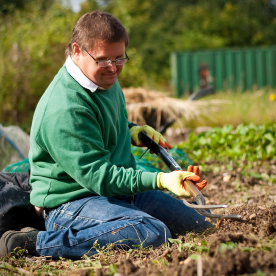 An Earthworker kneeling on the ground in the gardens, holding a shovel and wearing gardening gloves