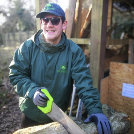An Earthworker sawing a log of wood outside, wearing a branded coat and cap, wearing protective gloves