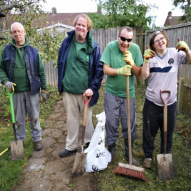 Four Earthworkers standing in an older person’s garden holding shovels and a broom