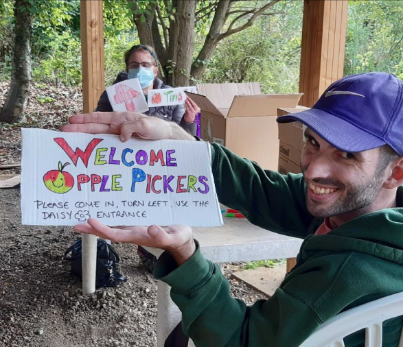An Earthworker sat at a table in the gardens, holding up a hand-written sign which says “Welcome Apple Pickers”