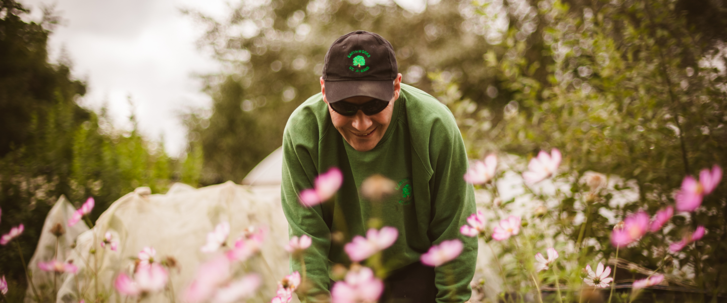 An Earthworker leaning down to flowers in a meadow, wearing a branded cap and sunglasses