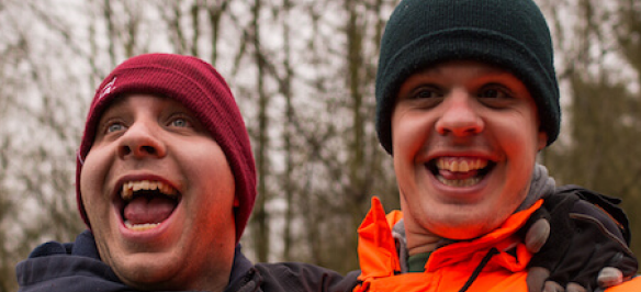 A volunteer and an Earthworker smilling outdoors wearing beanie hats and coats, with forest trees in the background